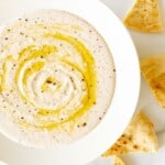 A white bowl filled with homemade white bean dip, drizzled with olive oil, pita triangles to the side.