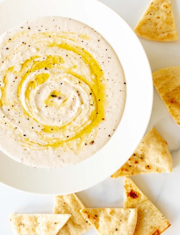 A white bowl filled with homemade white bean dip, drizzled with olive oil, pita triangles to the side.