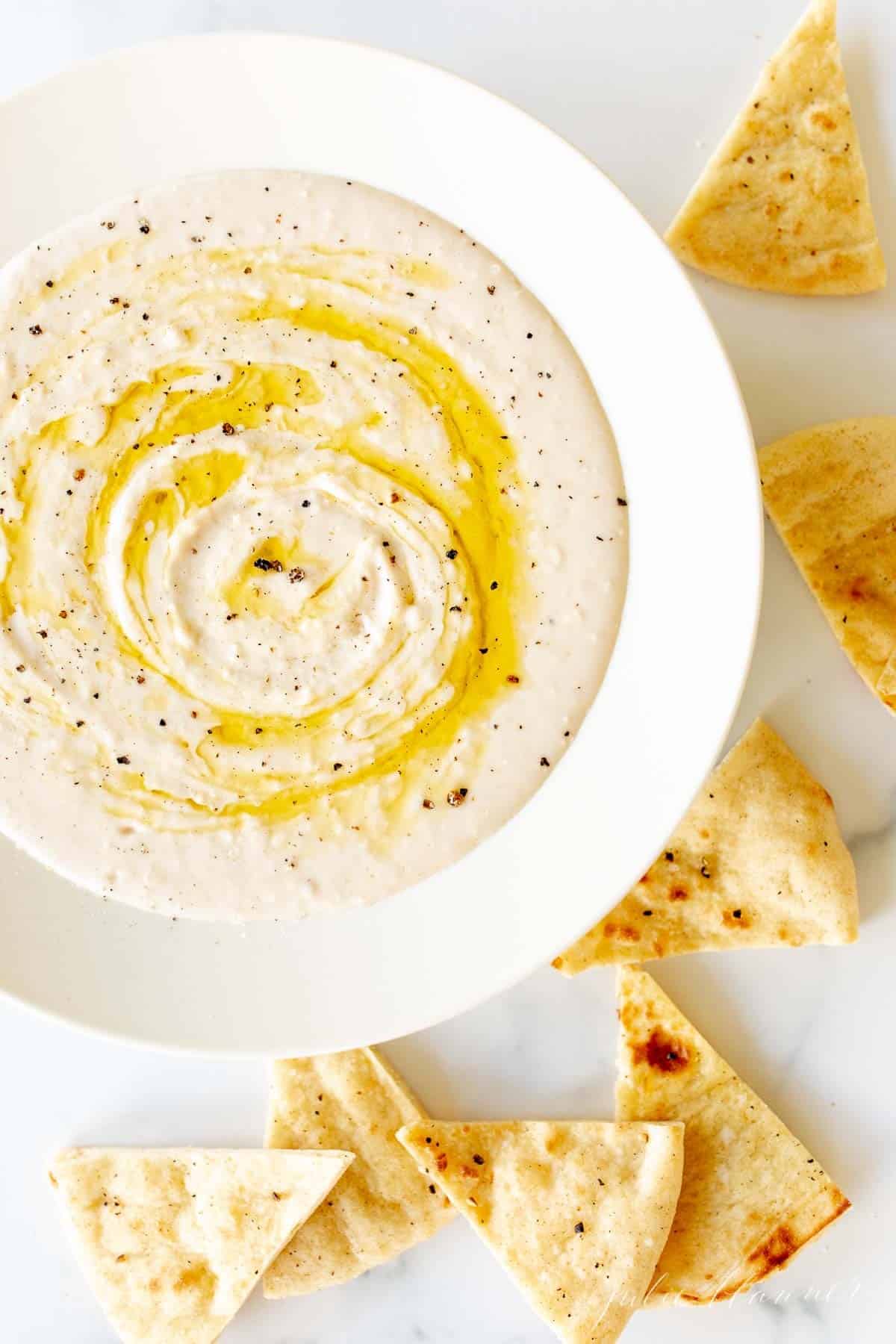 A white bowl filled with homemade white bean dip, drizzled with olive oil, pita triangles to the side.