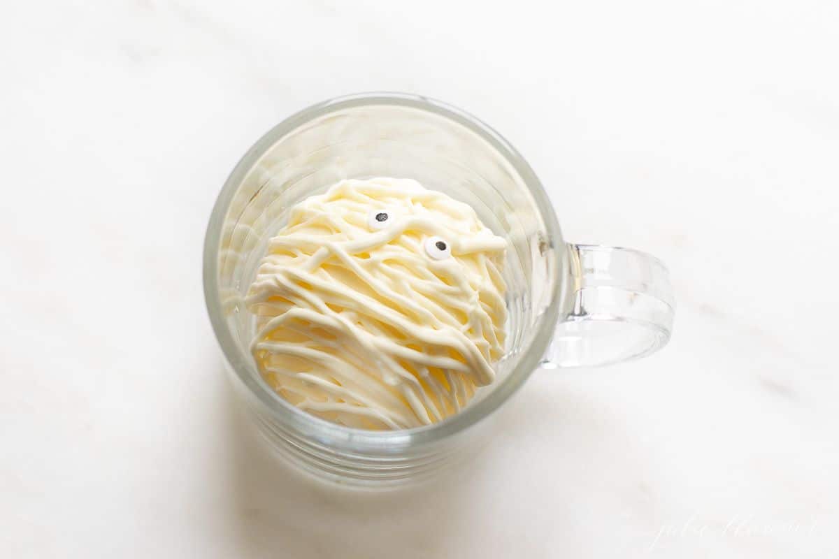 A white mummy Halloween hot chocolate bomb in a clear glass mug on a marble surface.
