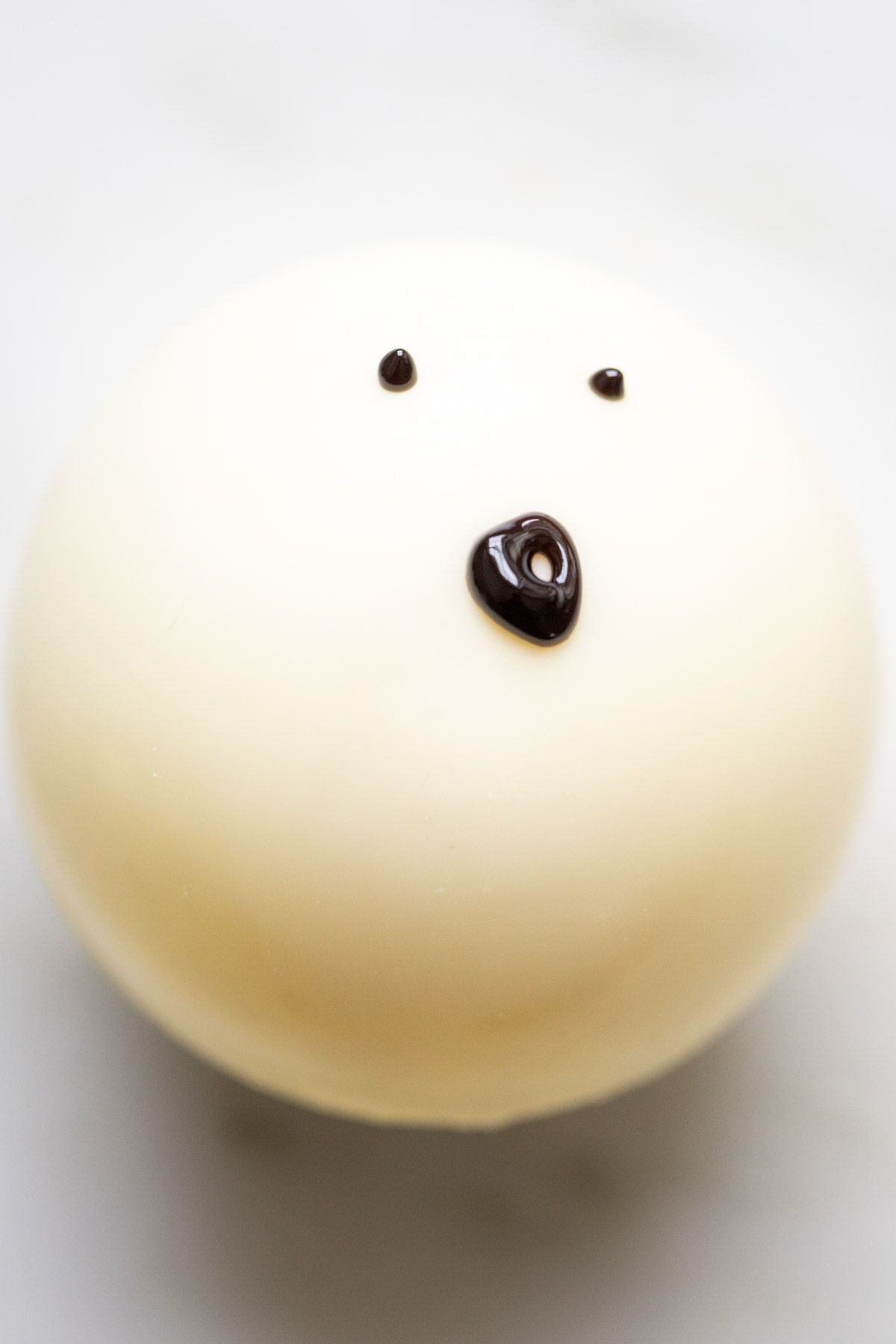 A white chocolate hot cocoa bomb, decorated as a ghost face.