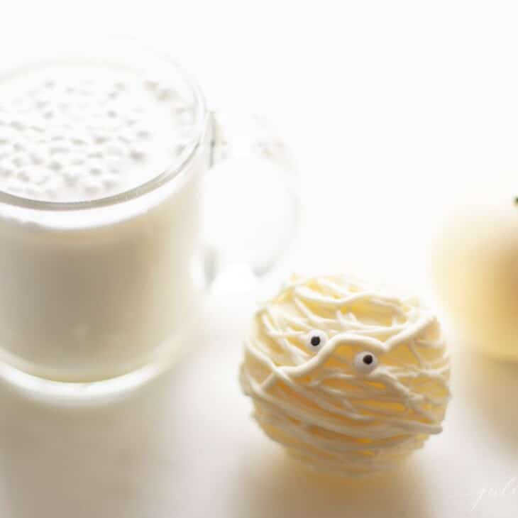 A clear glass mug of white chocolate hot cocoa and a Halloween hot chocolate bomb.