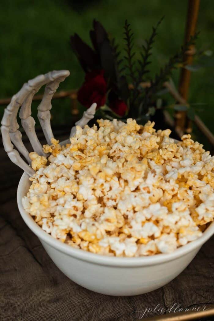 A white bowl full of popcorn with a skeleton hand reaching out for Halloween decorations.