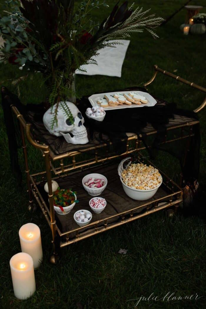 Halloween outdoor movie night snacks set up on a gold bar cart, flowers in the background.