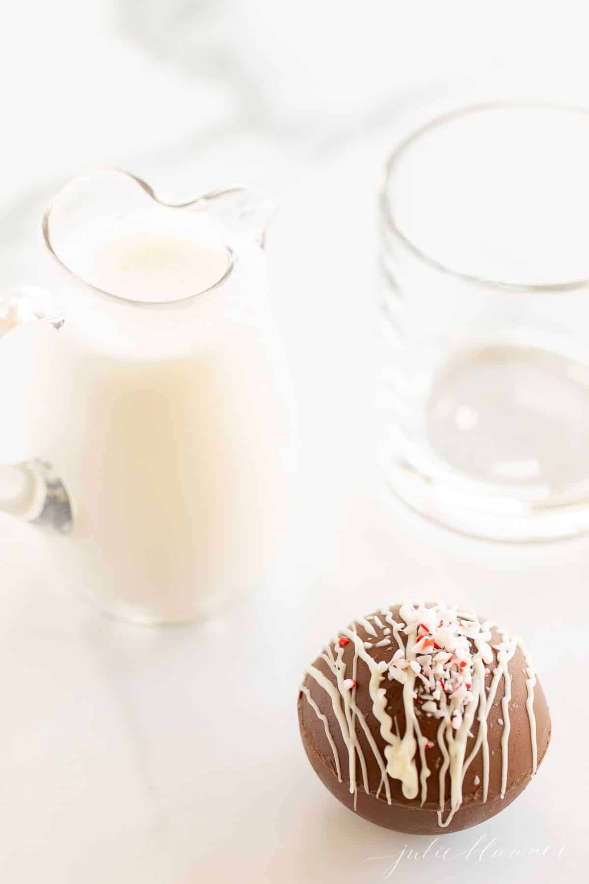 A clear glass pitcher of warm milk, clear mug in background, and a hot chocolate bomb.