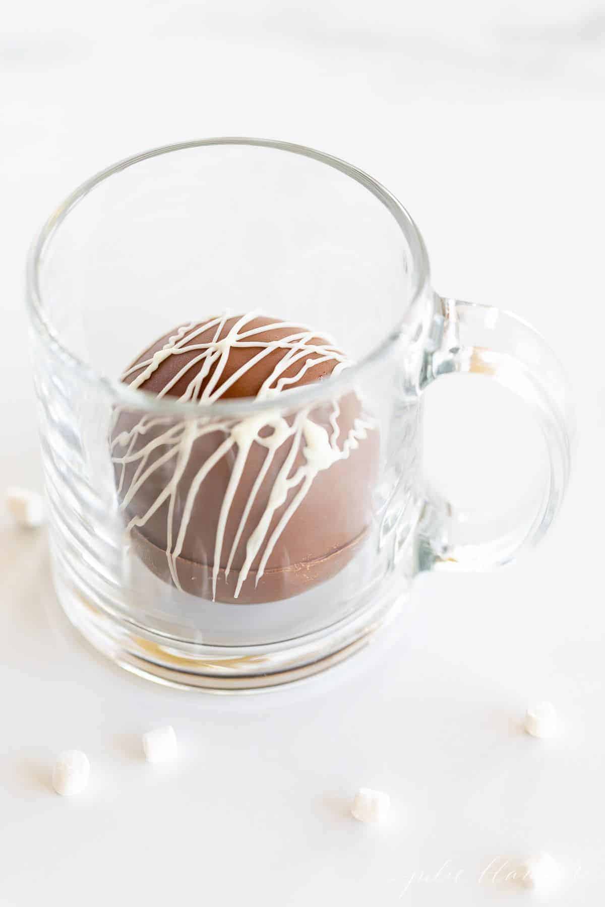 A clear glass mug with a single hot chocolate bomb inside, marshmallows surrounding mug on a marble surface.