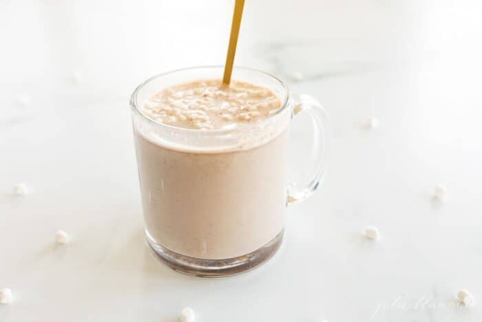 A clear glass mug of hot chocolate with a gold spoon sticking out.