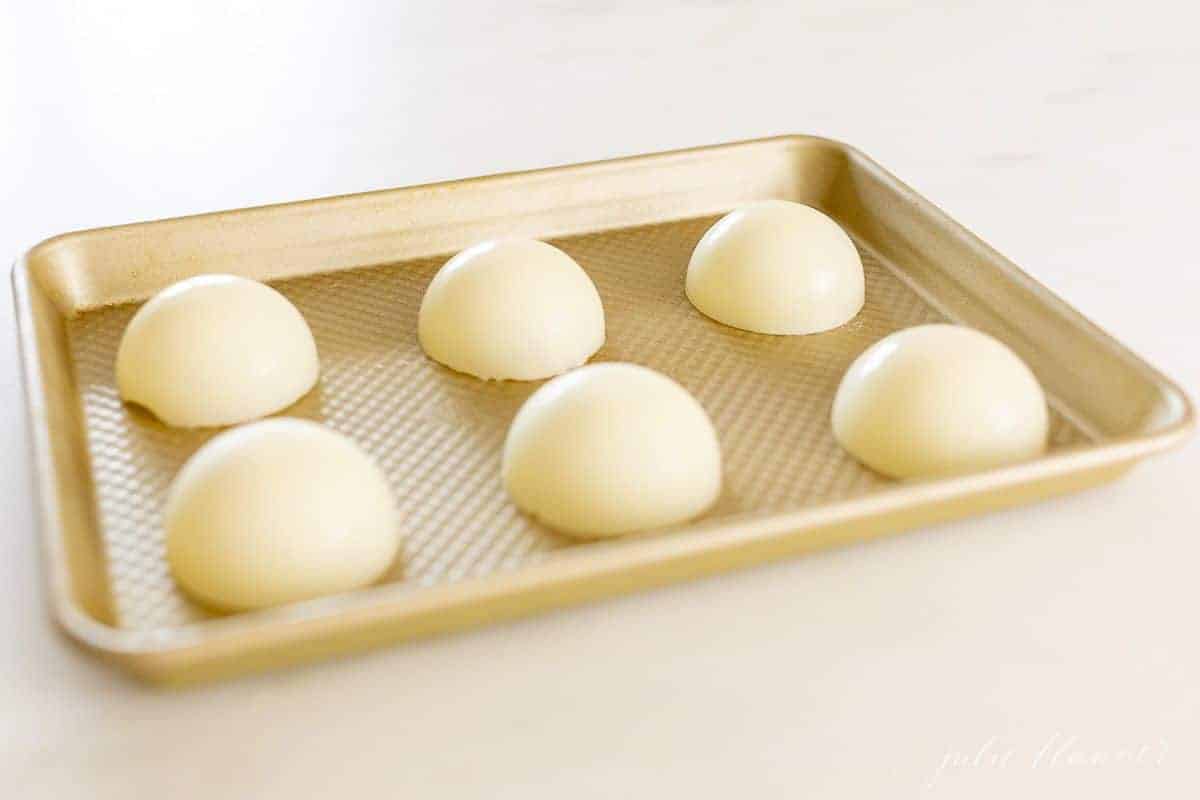 A gold baking sheet topped with white chocolate spheres for Halloween hot chocolate bombs.
