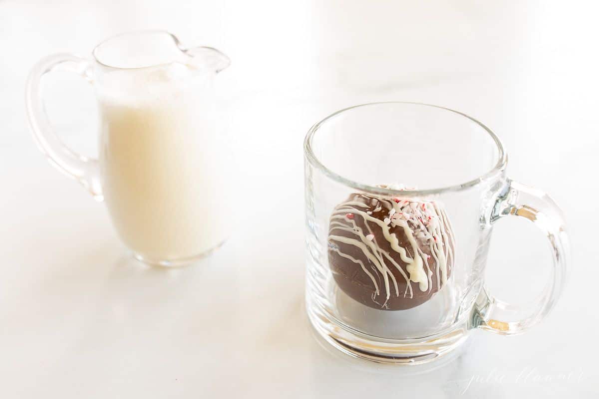 A clear glass pitcher of warm milk, clear mug in background, and a hot chocolate bomb.