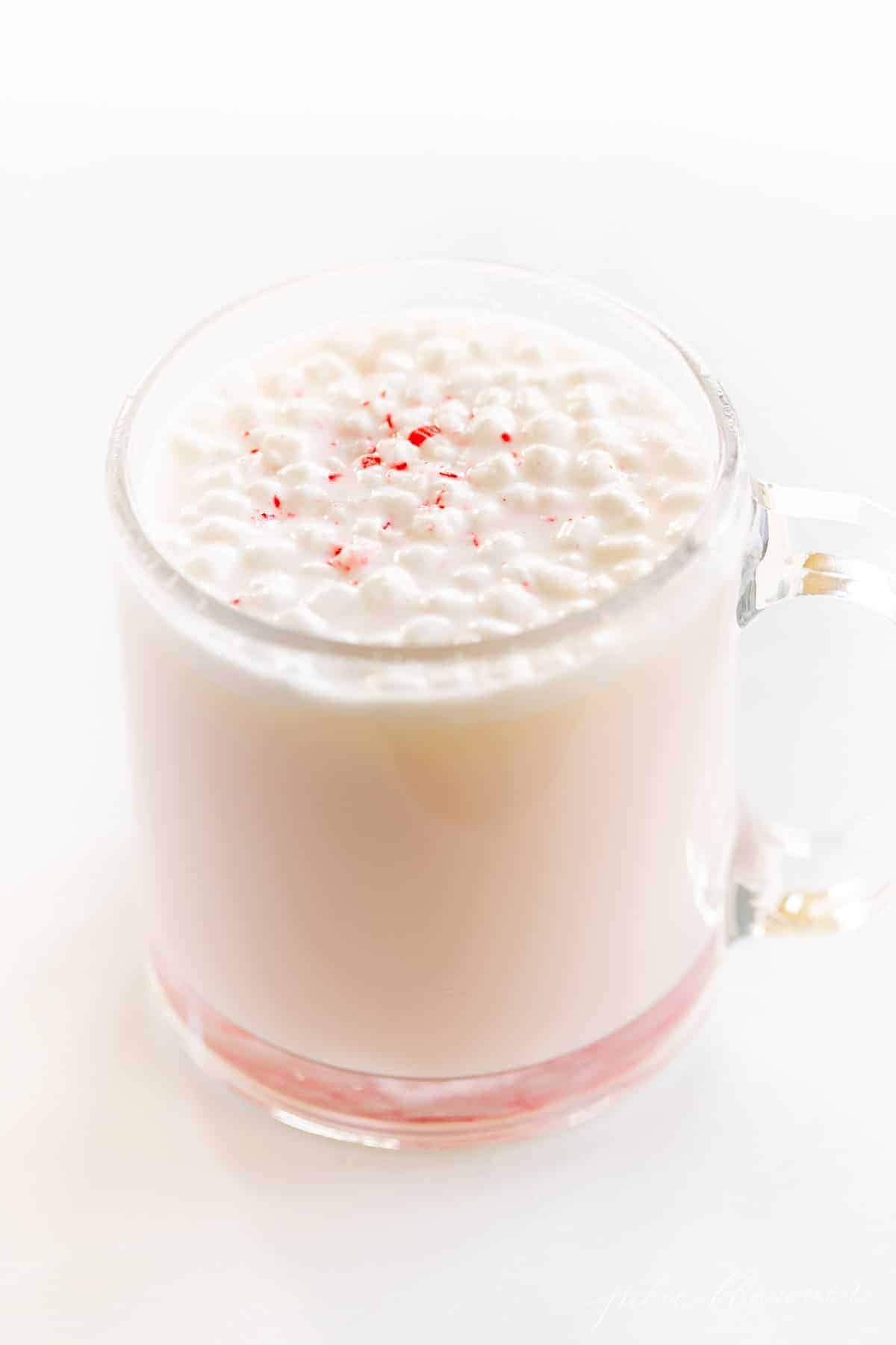 A clear glass mug of peppermint hot chocolate, topped with tiny marshmallows and crushed peppermint.