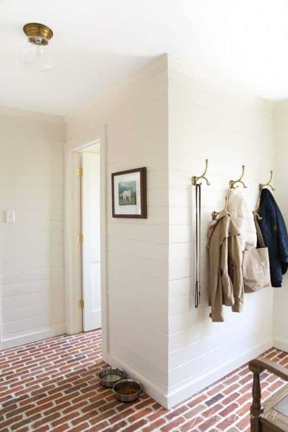 A white mudroom with brick floors and brass hooks