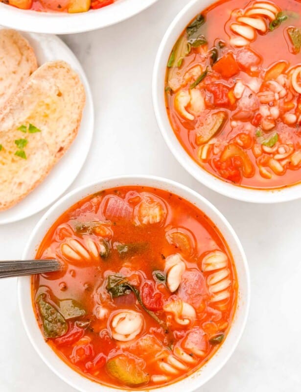 A white bowl of minestrone soup, another bowl in the background with herbs and a slice of bread on the surface.