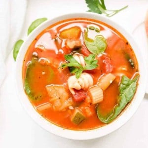 A white bowl of minestrone soup with herbs and a slice of bread on the surface.