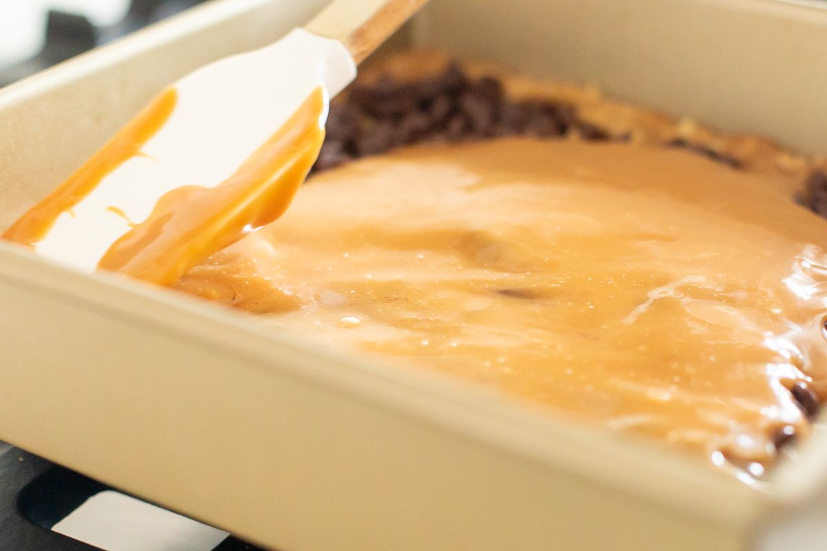 A spatula spreading melted caramel onto cookie bars in a gold pan
