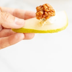 A hand holding a slice of pear topped with a walnut and blue cheese.