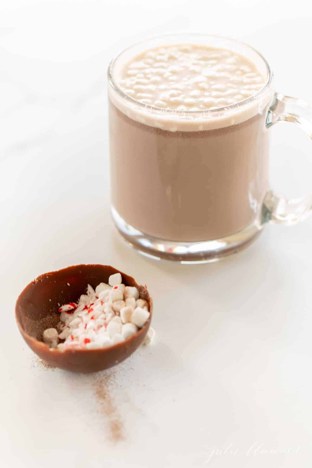 A clear glass mug full of hot chocolate with half a sphere of a hot chocolate bomb in the foreground.