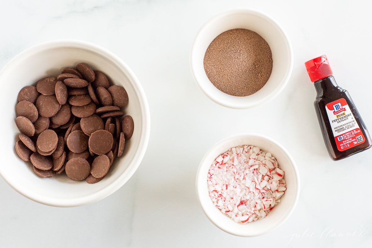 Peppermint hot chocolate bombs and chocolate ingredients in bowls on a marble countertop.