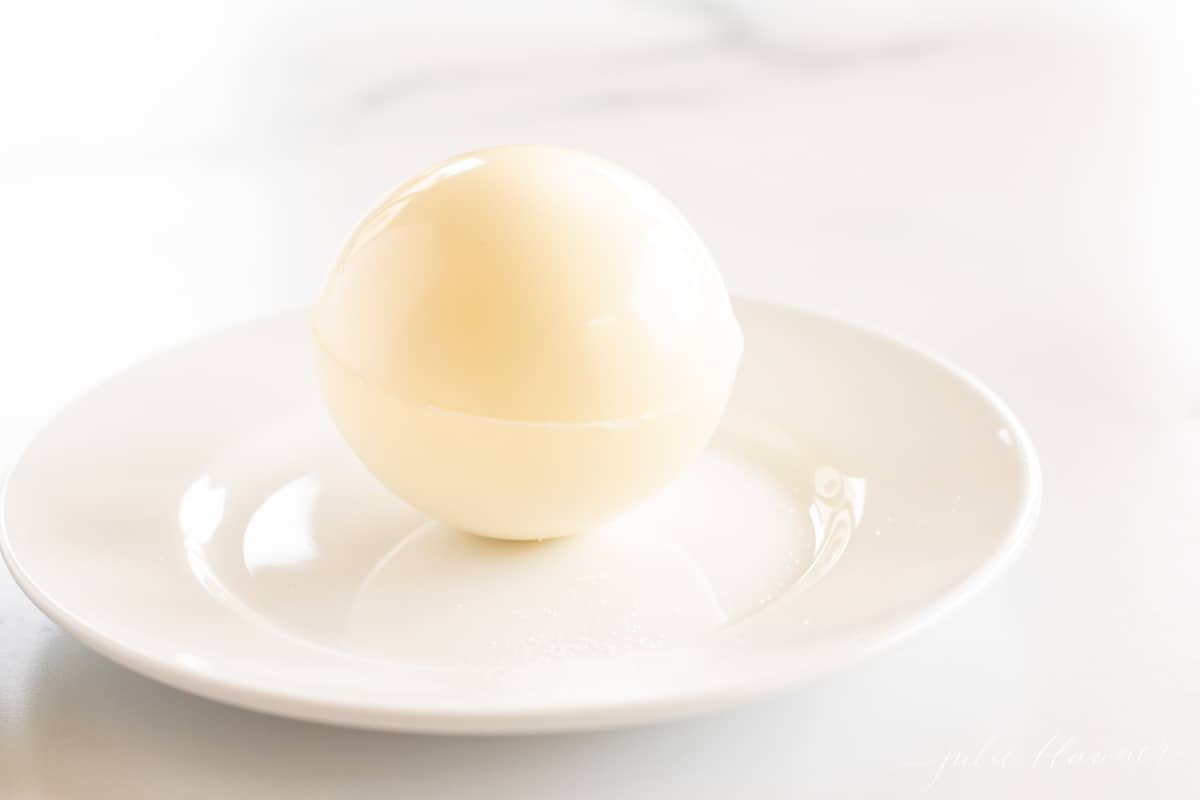 A white chocolate hot chocolate bomb on a white plate on a marble surfacde.