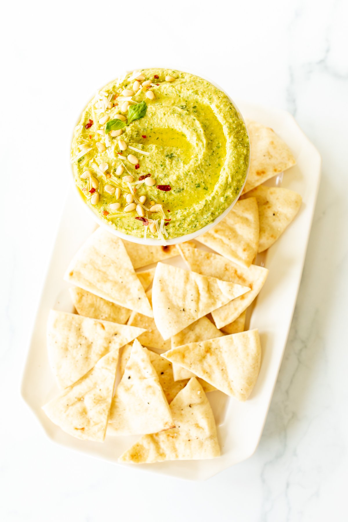 Pesto hummus in a white bowl surrounded by pita bread.