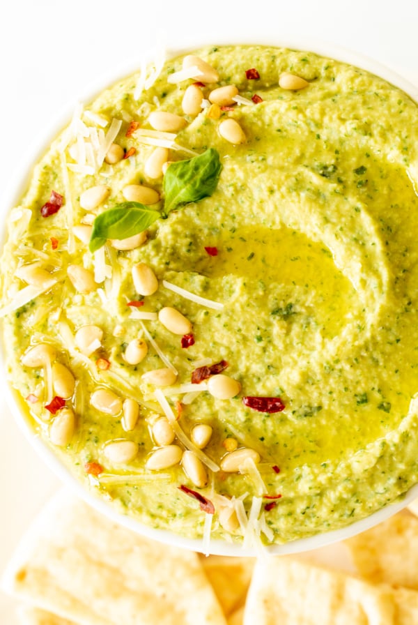 A white bowl full of basil pesto hummus, topped with pine nuts and parmesan.