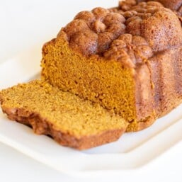 A white platter featuring a loaf of pumpkin bread with a slice cut at the front.