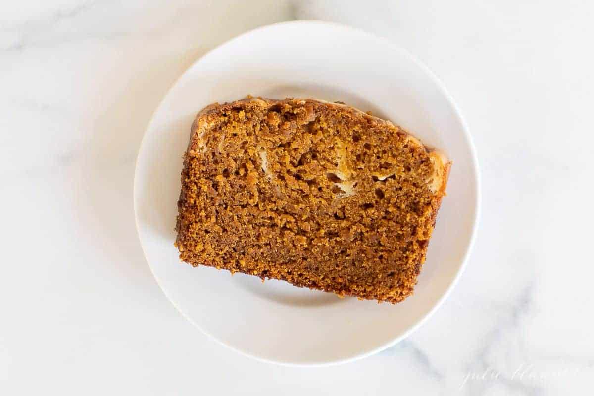 A marble surface with a small white plate with a slice of pumpkin cheesecake bread.
