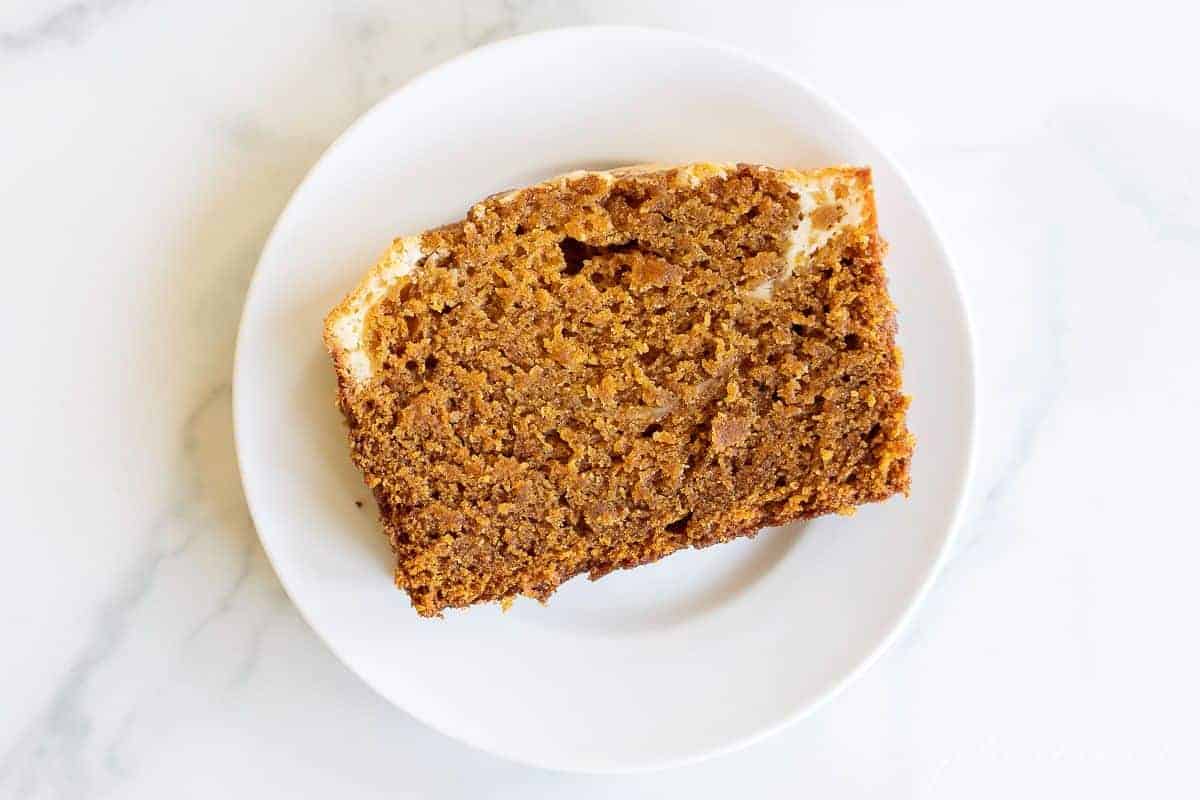 A marble surface with a small white plate with a slice of pumpkin cheesecake bread.