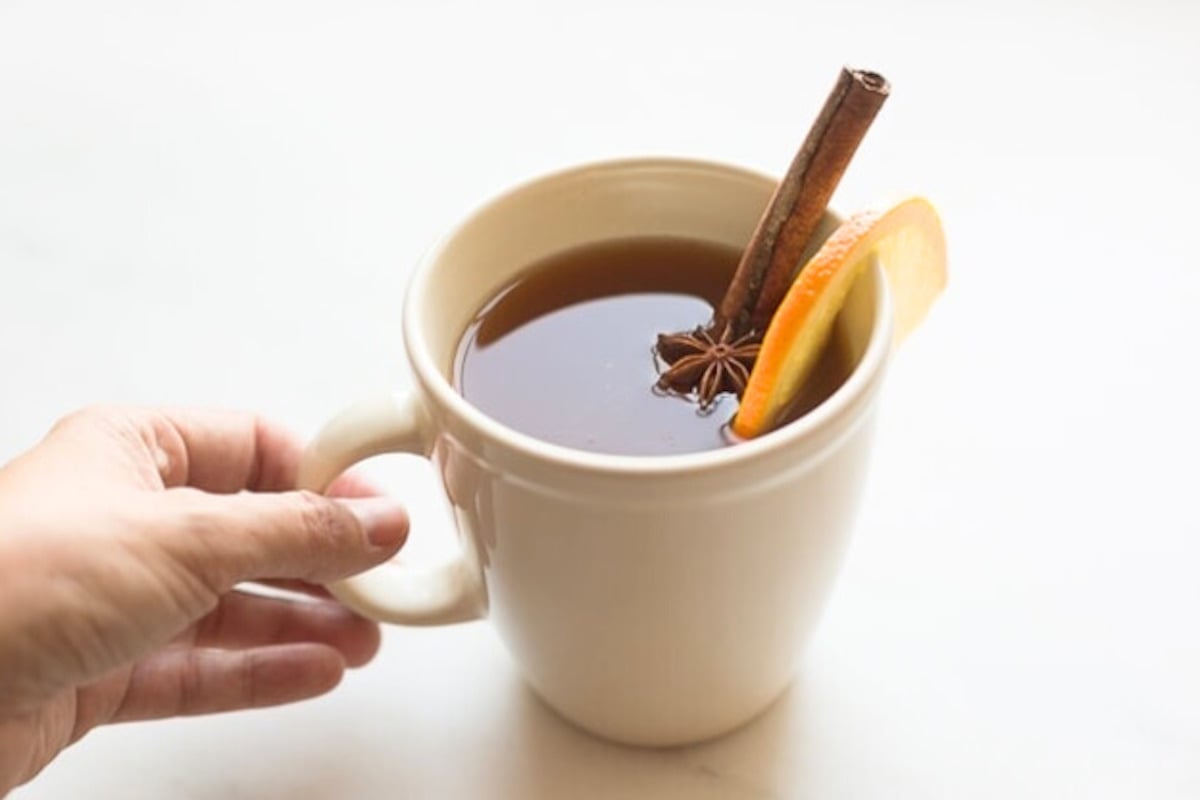 A hand holds a white mug filled with a dark liquid, garnished with a slice of orange, a cinnamon stick, and a star anise—perfect for enjoying spiked apple cider.