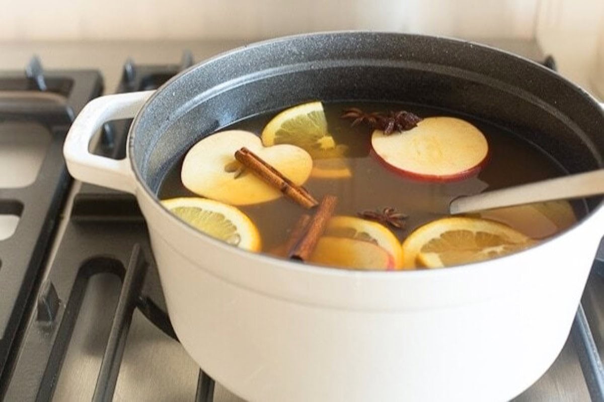 A white pot on a stove filled with hot spiked apple cider, garnished with slices of apple and lemon, cinnamon sticks, and star anise.