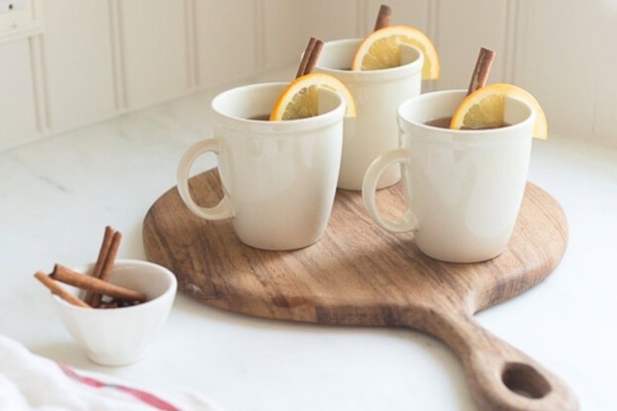 Three white mugs with lemon slices and cinnamon sticks on a round wooden serving board, next to a small bowl of cinnamon sticks, all placed on a white countertop, perfect for serving spiked apple cider.