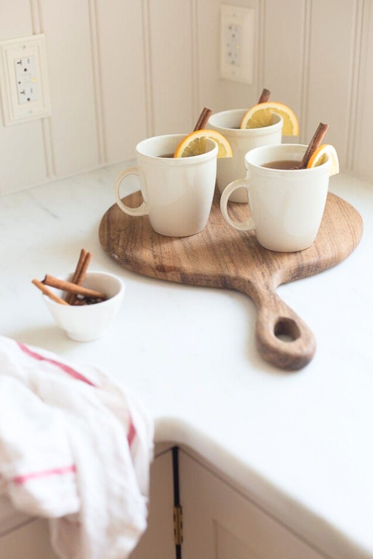 Three white mugs filled with spiked apple cider, each garnished with a cinnamon stick and a lemon slice, placed on a wooden board. A small bowl with extra cinnamon sticks and a cloth are beside them.