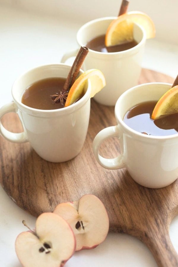 Three white mugs filled with a brown liquid, garnished with orange slices and cinnamon sticks, are placed on a wooden board beside apple slices—perfect for enjoying spiked apple cider.