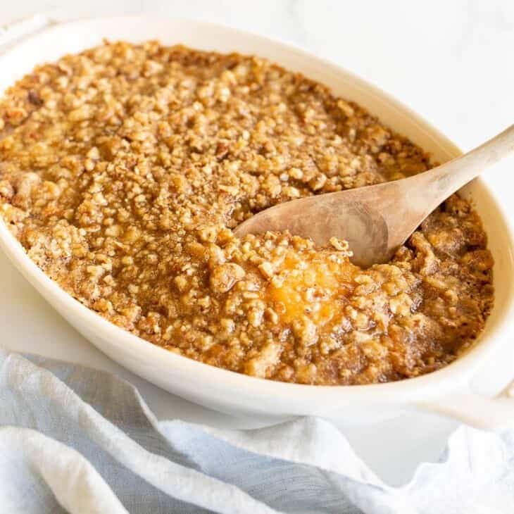 A white oval baking dish filled with a sweet potato casserole, spoon lifting out a serving.