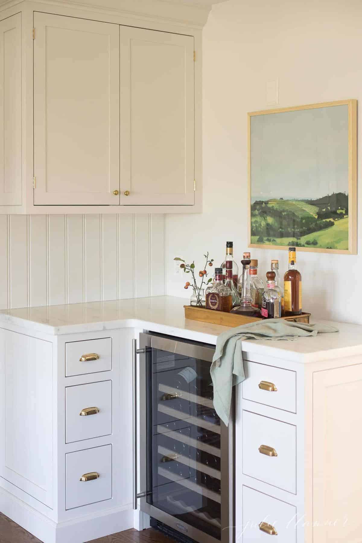 A corner of a white kitchen with a small bar tray set up with liquor over a wine refrigerator.