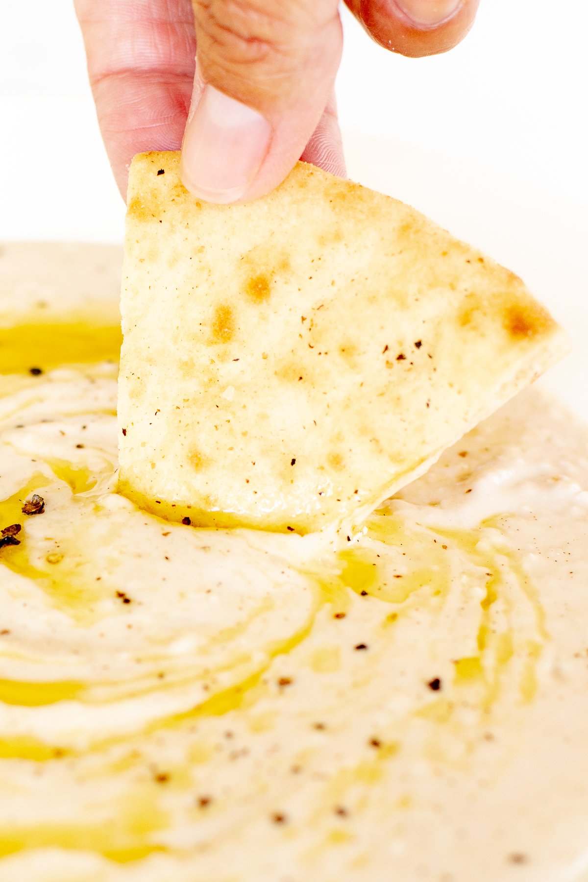 A hand dipping a triangle of pita bread into a bowl of white bean dip.