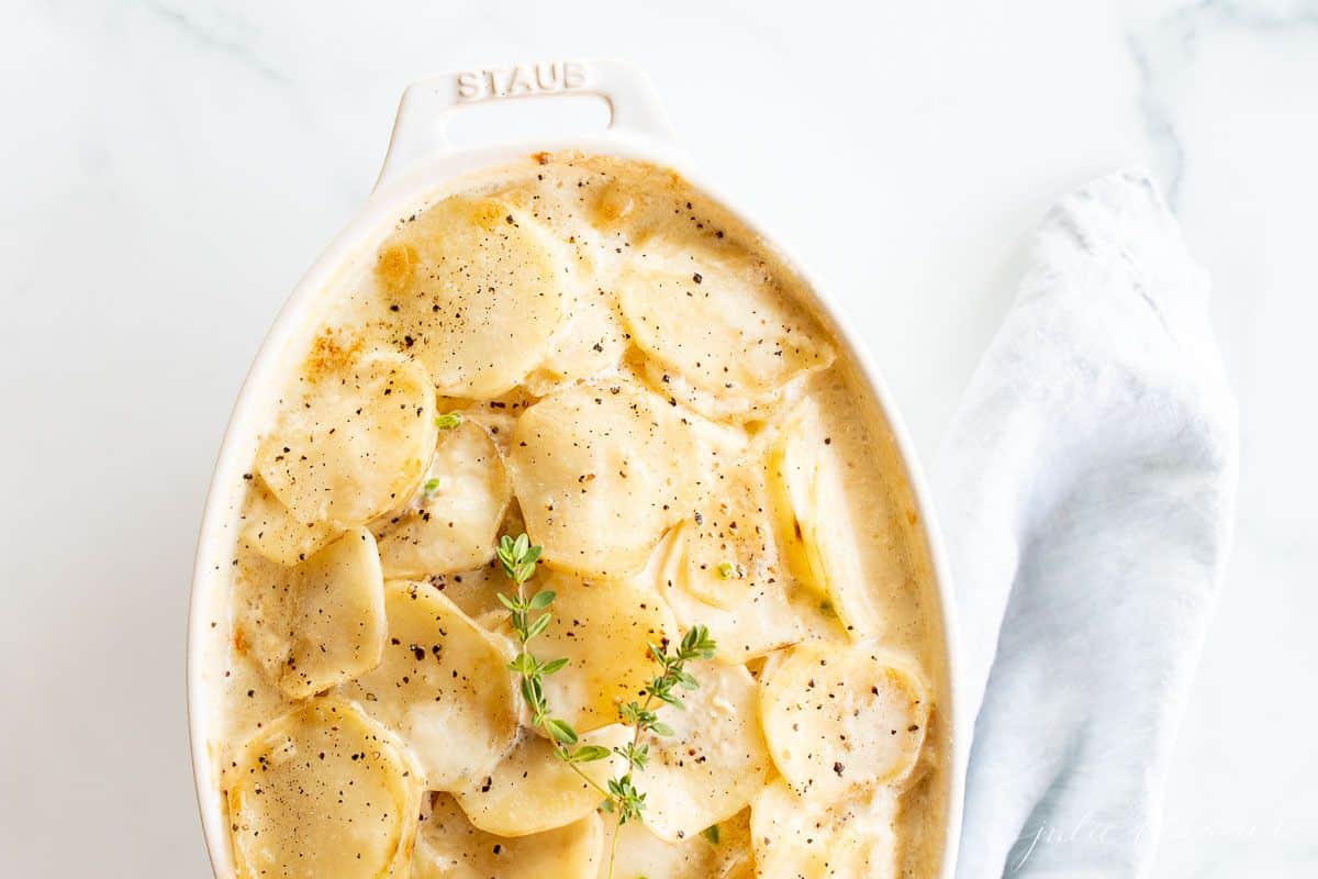 A white oval Staub baker filled with Gratin Dauphinoise potato bake, blue linen napkin to the side.