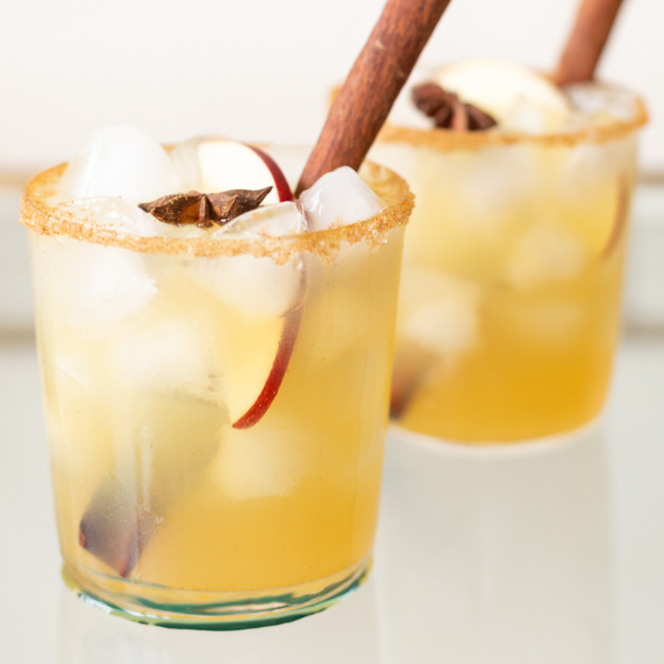 Clear glasses with apple cider margaritas, rimmed in sugar and garnished with a cinnamon stick on a marble surface.