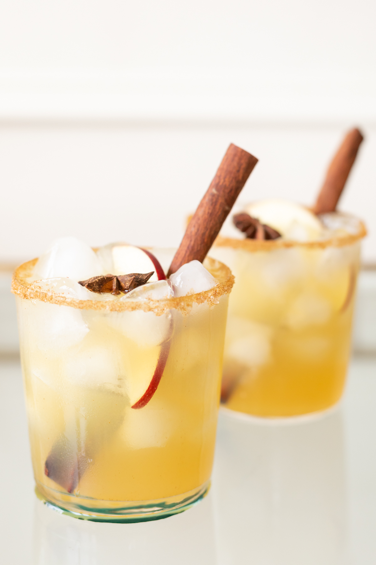 Clear glasses with apple cider margaritas, rimmed in sugar and garnished with a cinnamon stick on a marble surface.