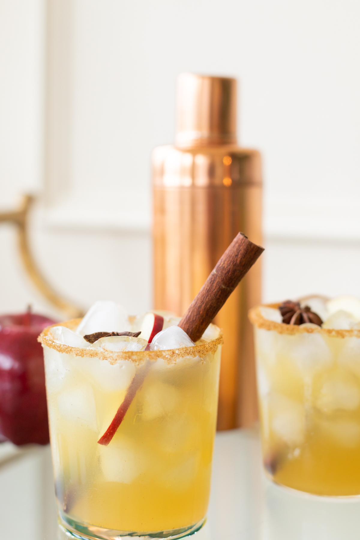 Clear glasses with apple cider margaritas, rimmed in sugar and garnished with a cinnamon stick with a copper shaker in the background.