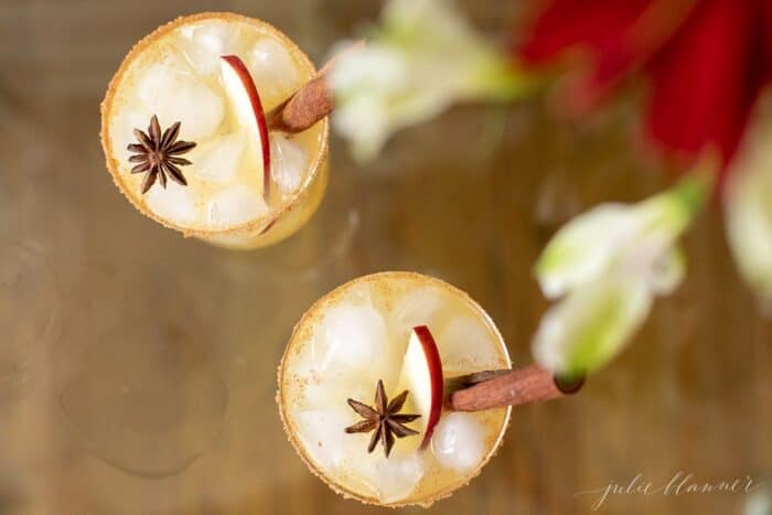 Clear glasses with apple cider margaritas, rimmed in sugar and garnished with a cinnamon stick on a wood surface.