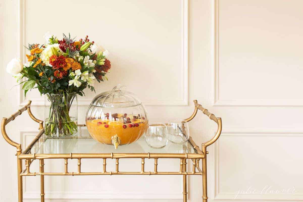 A glass bar cart with fall flowers in a vase, and a pumpkin drink dispenser filled with apple cider sangria.
