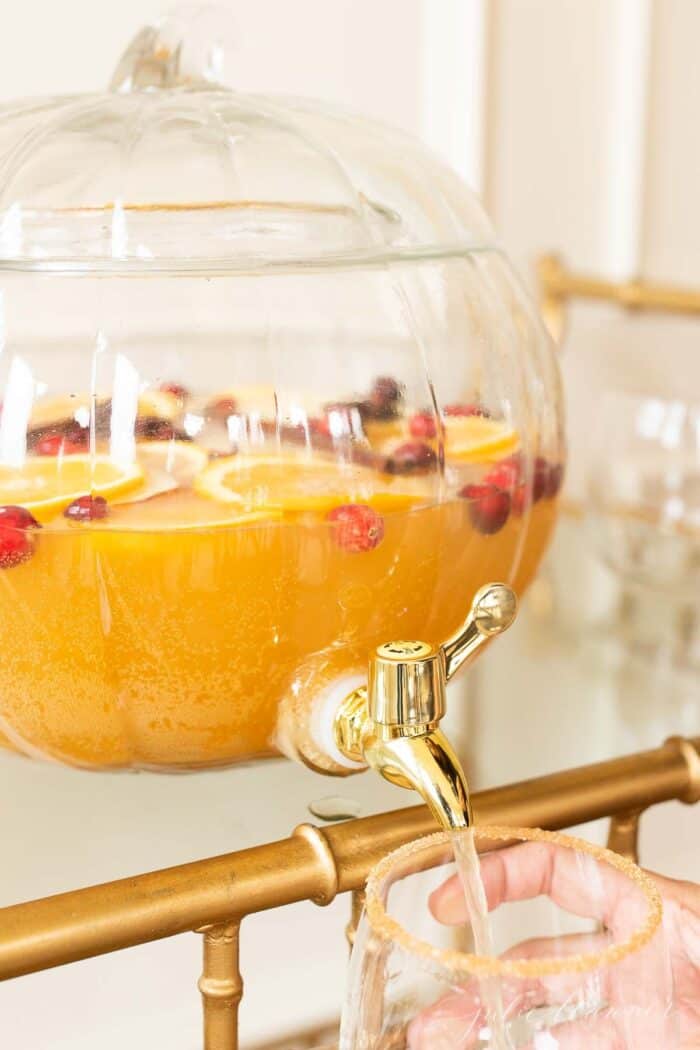 A clear glass pumpkin drink dispenser filled with an apple cider sangria recipe, dispensing fall sangria into a glass.