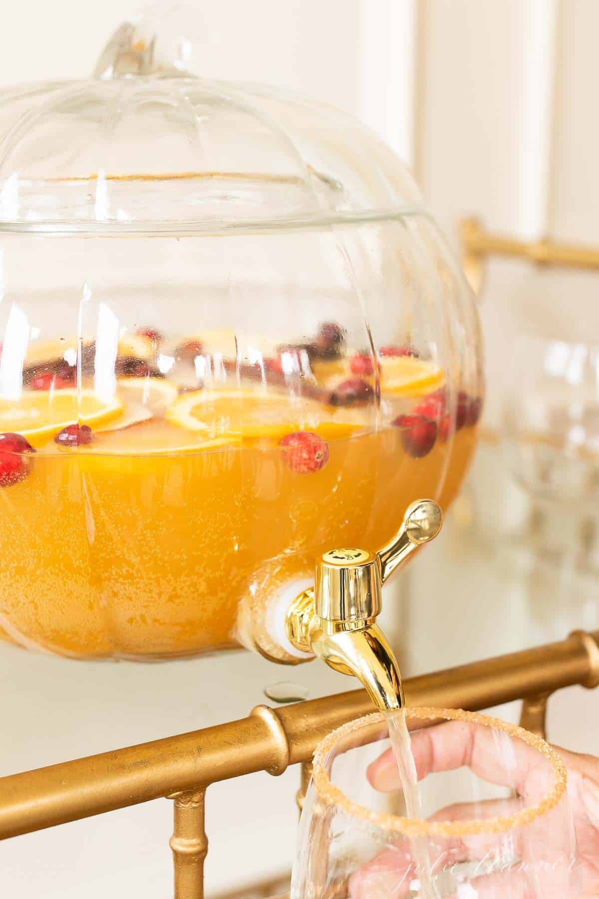 A clear glass pumpkin drink dispenser filled with an apple cider sangria recipe, dispensing fall sangria into a glass.