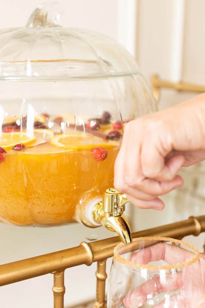 A clear glass pumpkin drink dispenser filled with an apple cider sangria recipe, dispensing fall sangria into a glass.
