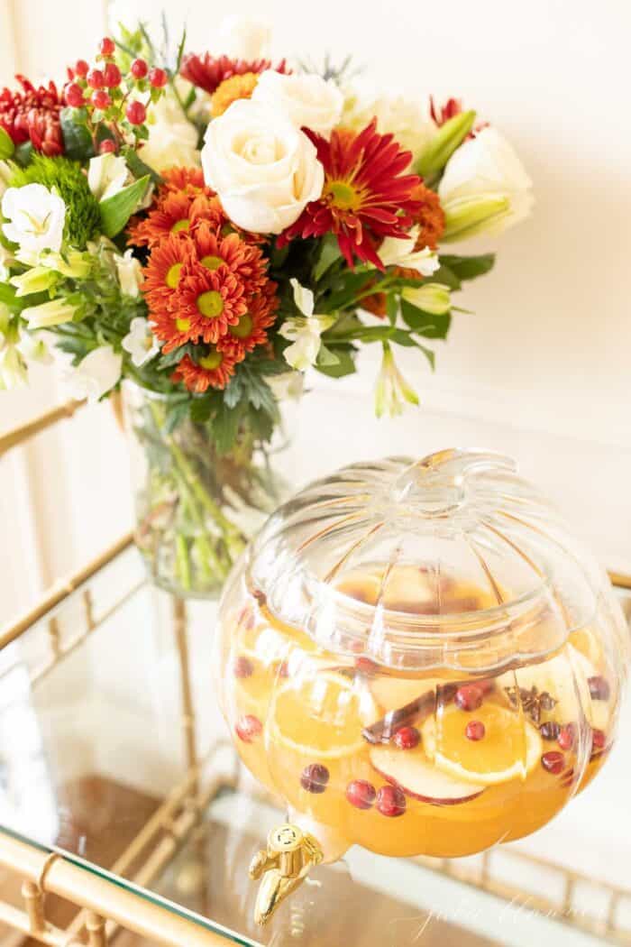 A clear glass pumpkin drink dispenser filled with apple cider sangria, flowers in the background.