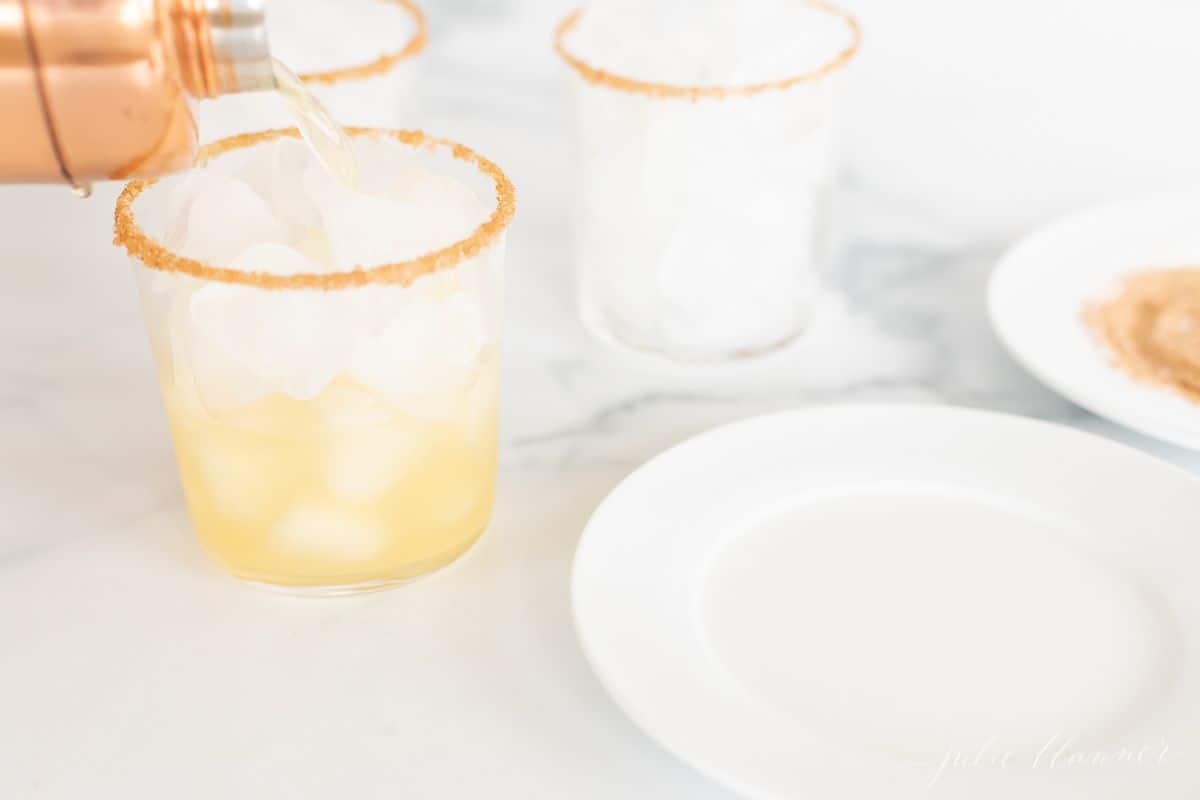 Three cocktail glasses with ice, a hand pouring an apple margarita into one from a copper mixer.