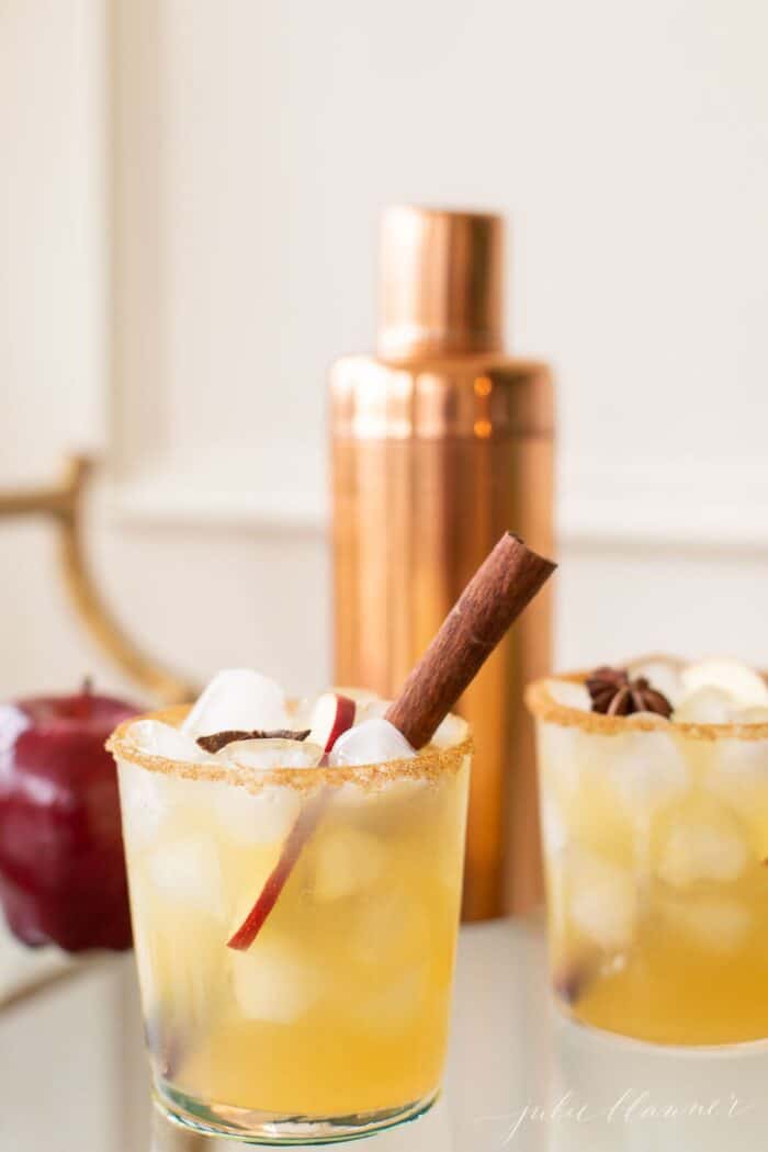 A copper cocktail shaker with clear glasses full of apple margaritas, cinnamon stick for garnish.