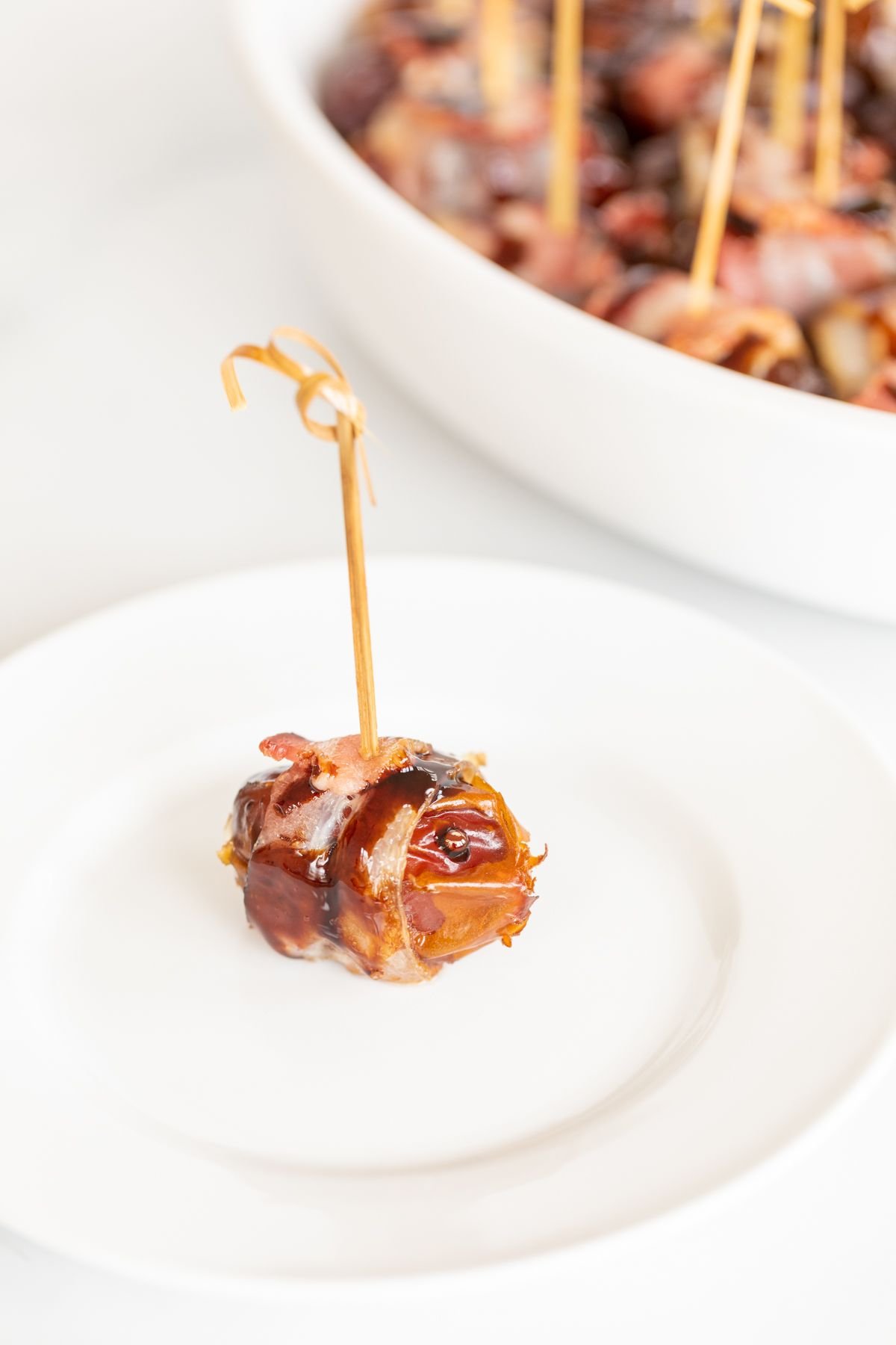 A single stuffed date wrapped in bacon on a white plate, with a dish full of them in the background.