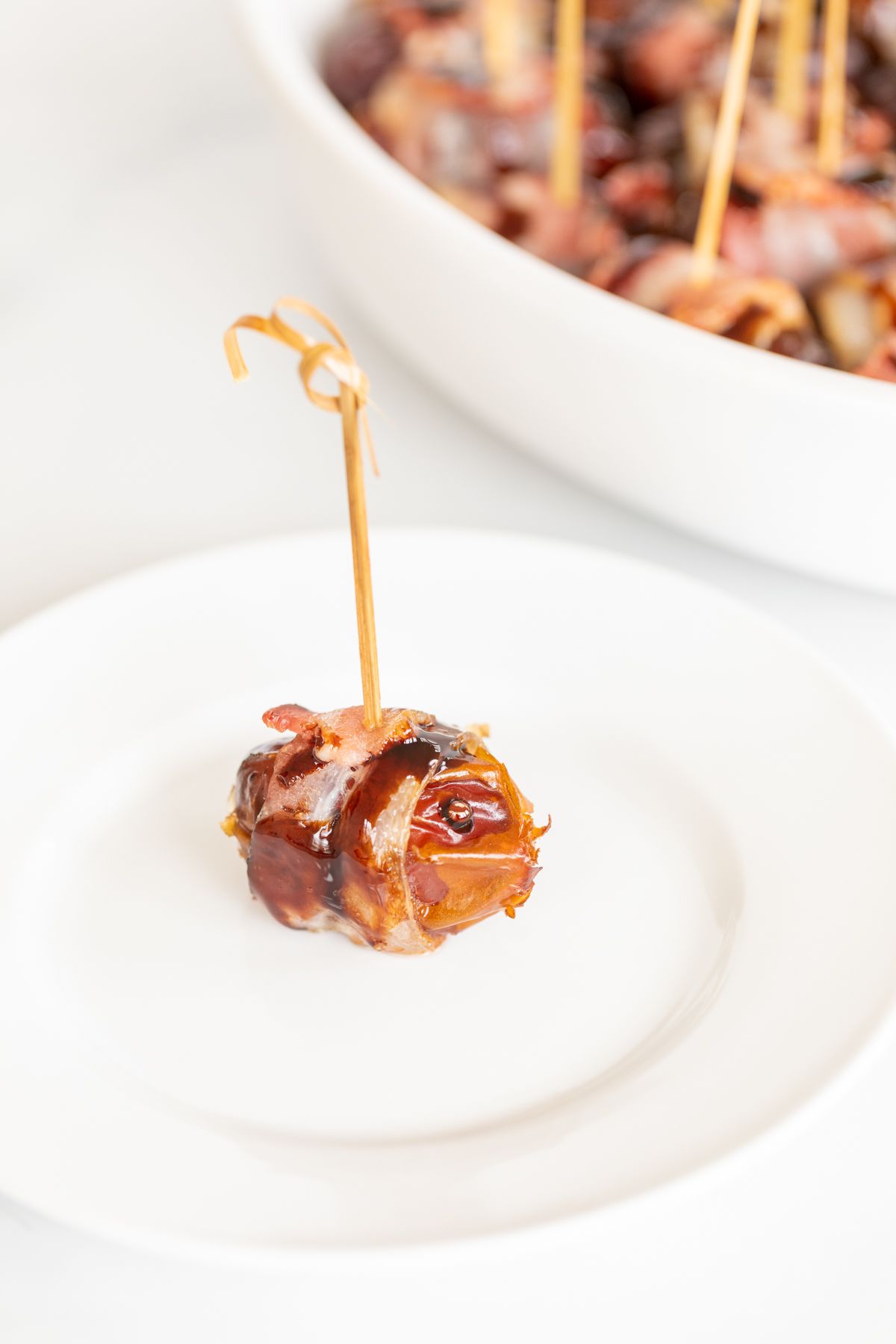 A single stuffed date wrapped in bacon on a white plate, with a dish full of them in the background.