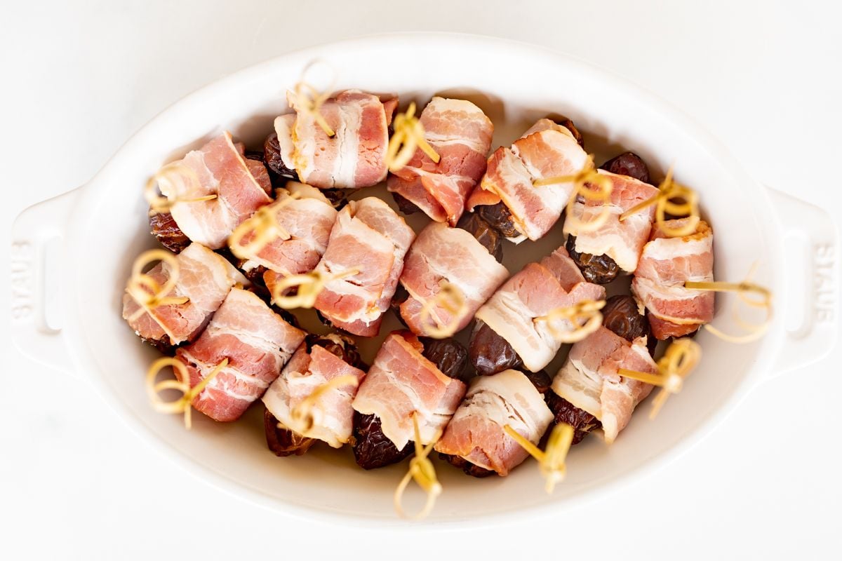 A white oval baking dish with bacon wrapped dates before baking, each has a toothpick for serving.